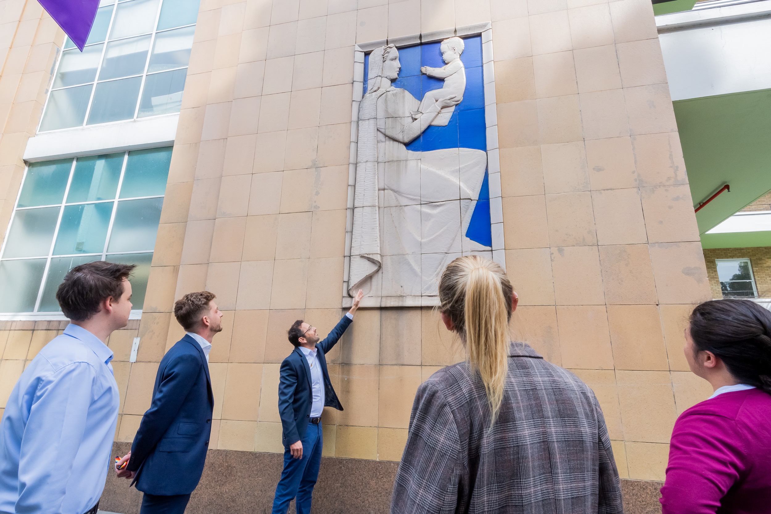 Participants look up as a tour guide points at historical exterior tiles crafted to depict a mother holding her newborn baby.