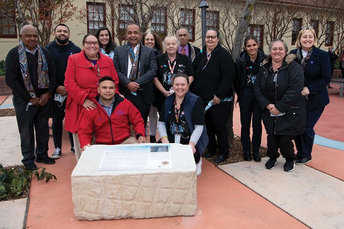 A group of people pose for a photo behind a plaque in a garden.