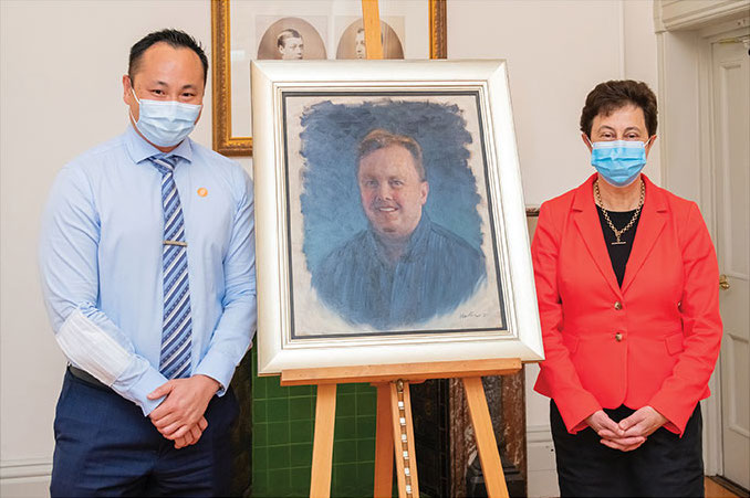 Two people pose for a photo by the painting of Robert Ogle.