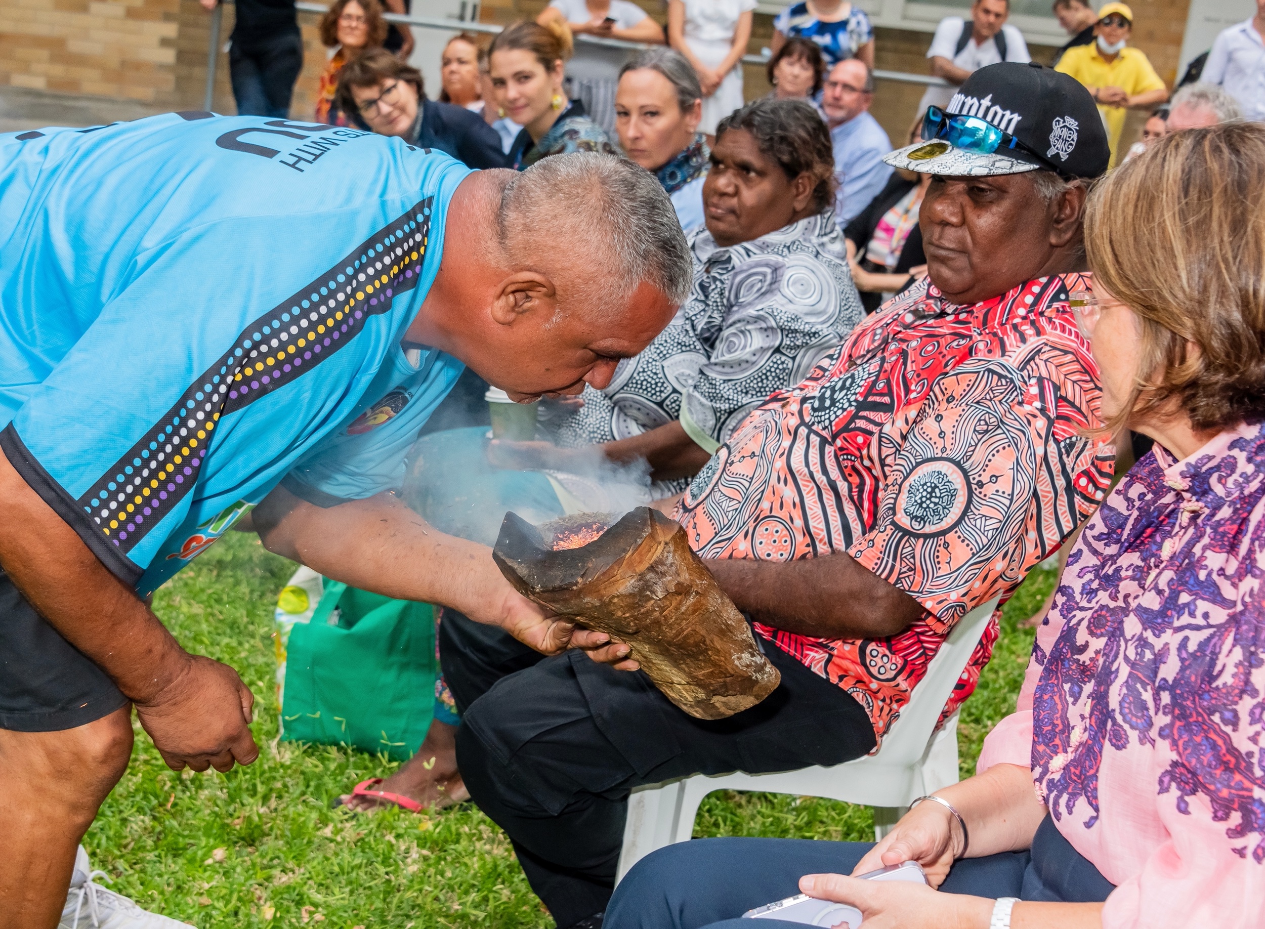 An Aboriginal man engages in a traditional smoking ceremony with a crowd of participants.