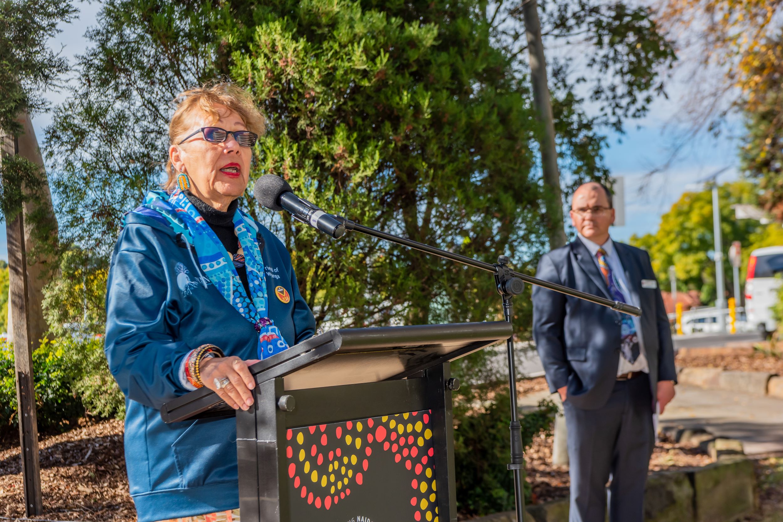 Woman talking at a lectern; man standing behind her