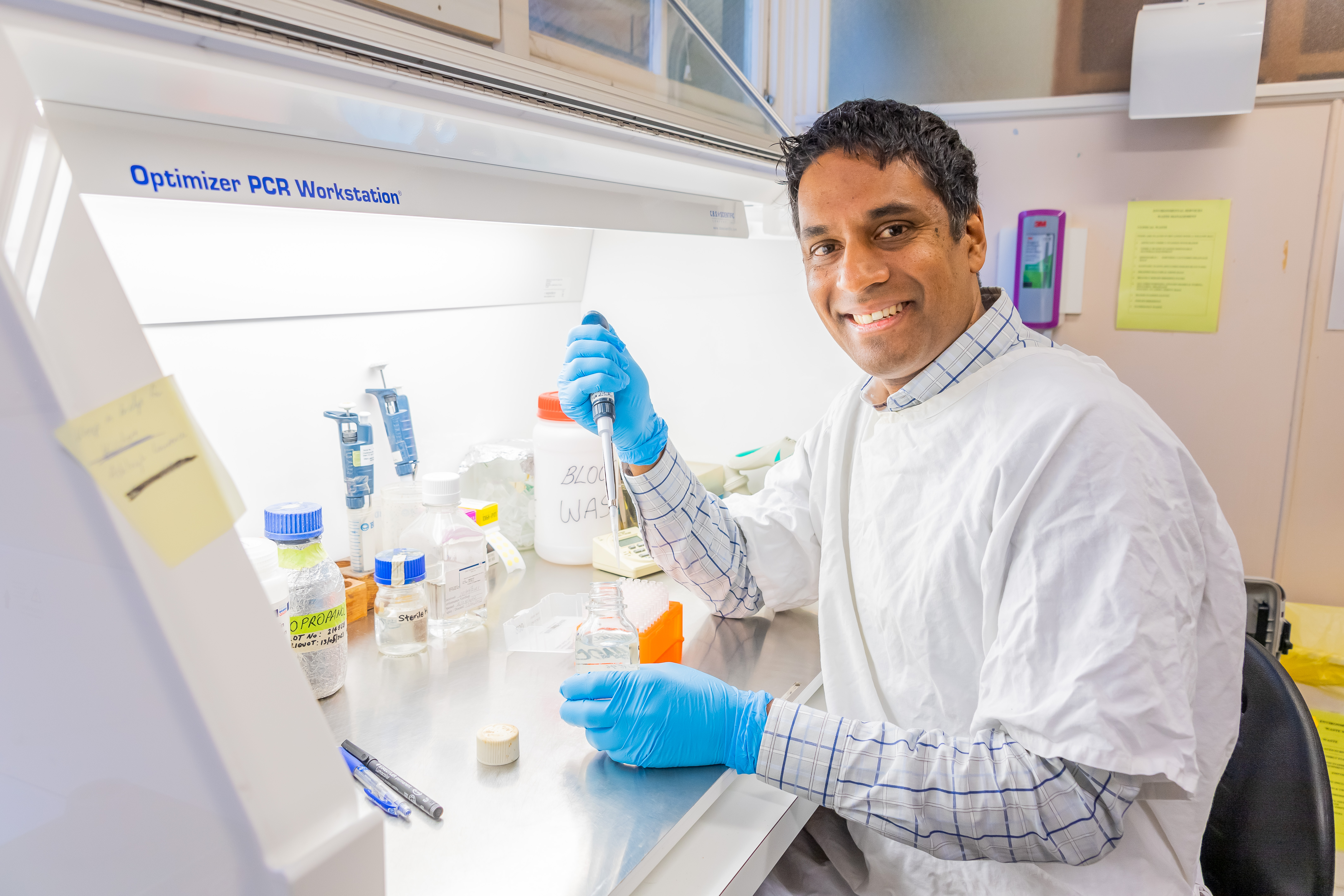 Dr Kishor Kumar is sitting at a lab desk, smiling at the camera. He wears a lab coat and is holding some lab equipment.