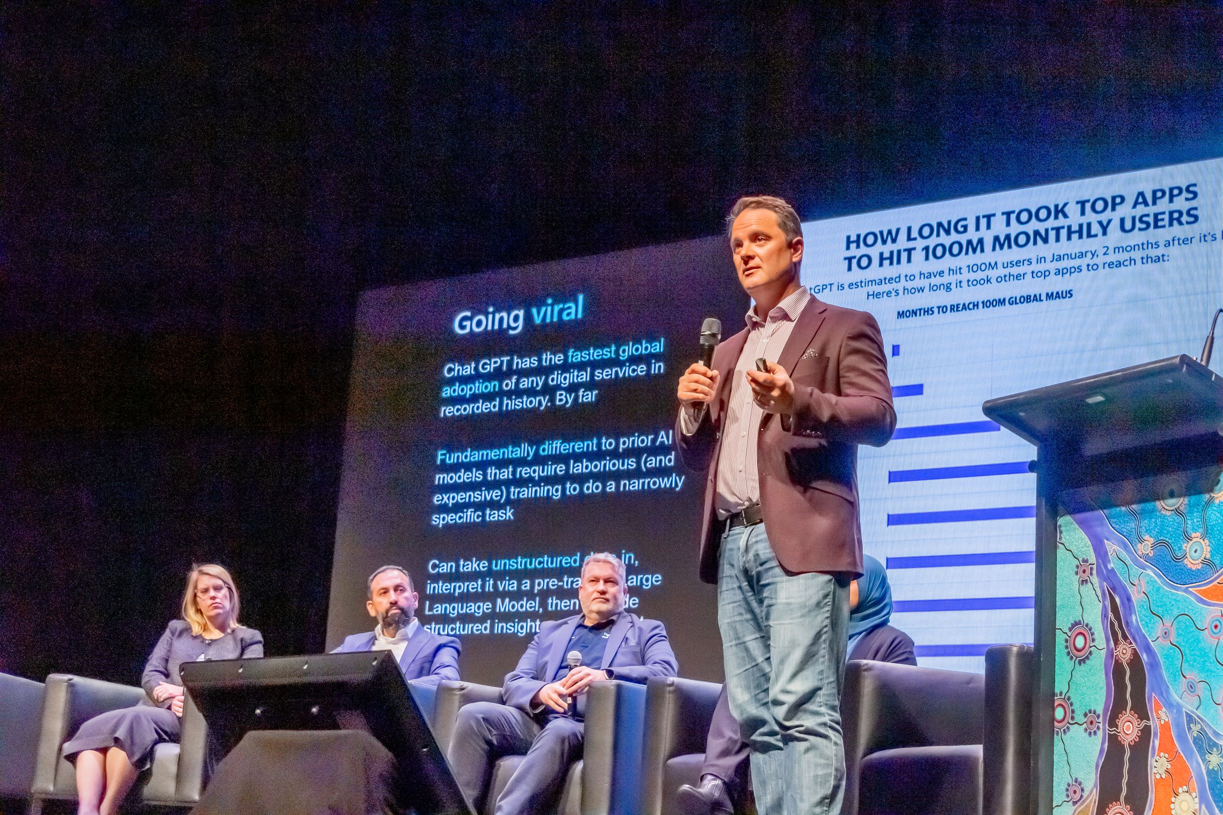 Man talking in front of a panel of people and a screen