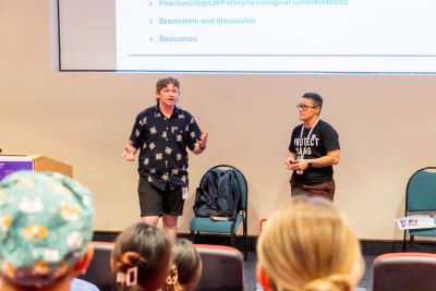 A man speaks to an auditorium with his arms held open, standing alongside a colleague.