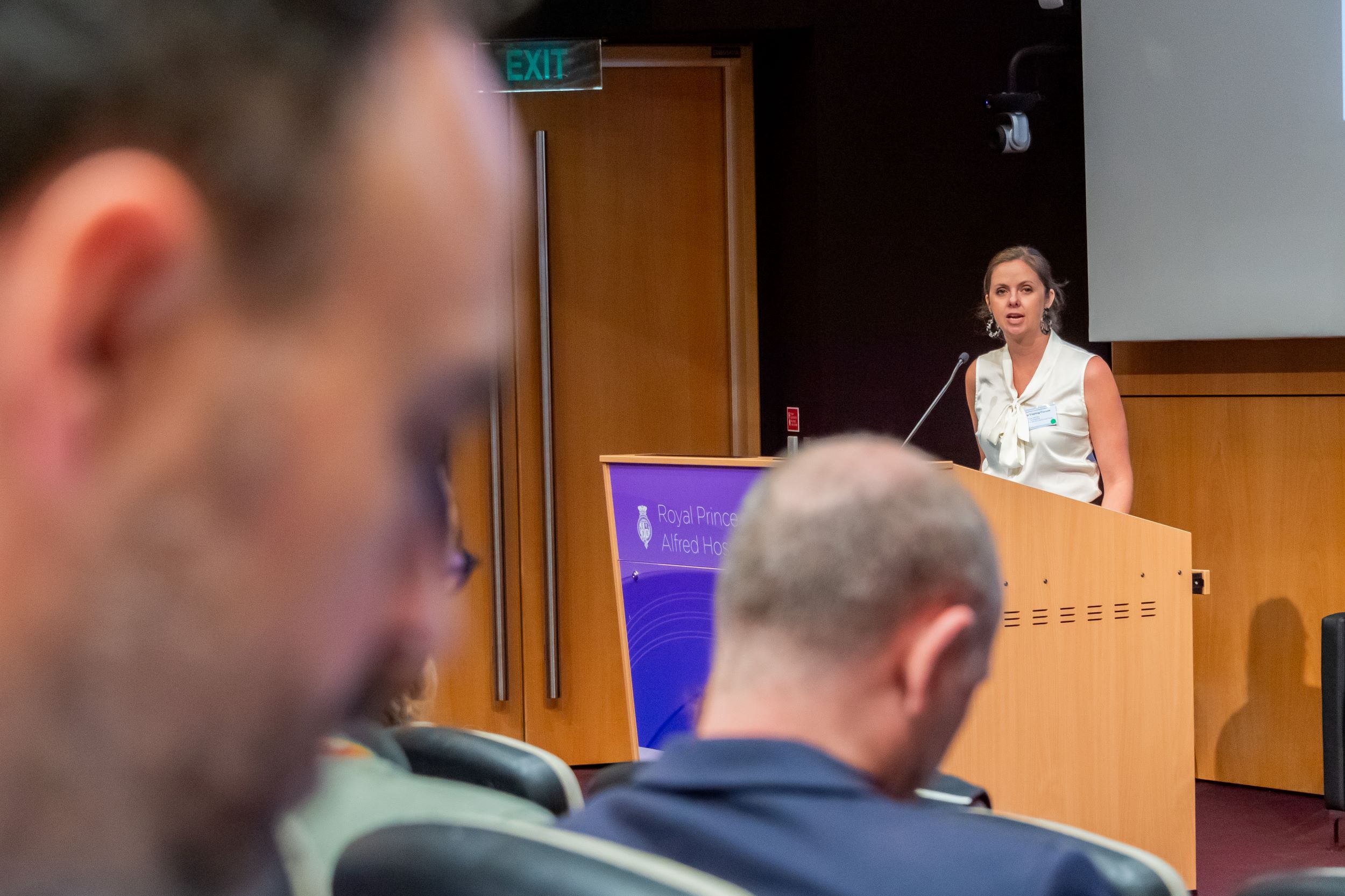 A woman is standing at a lecturn speaking to an auditorium crowd. In the foreground, several members of the audience are visible. 