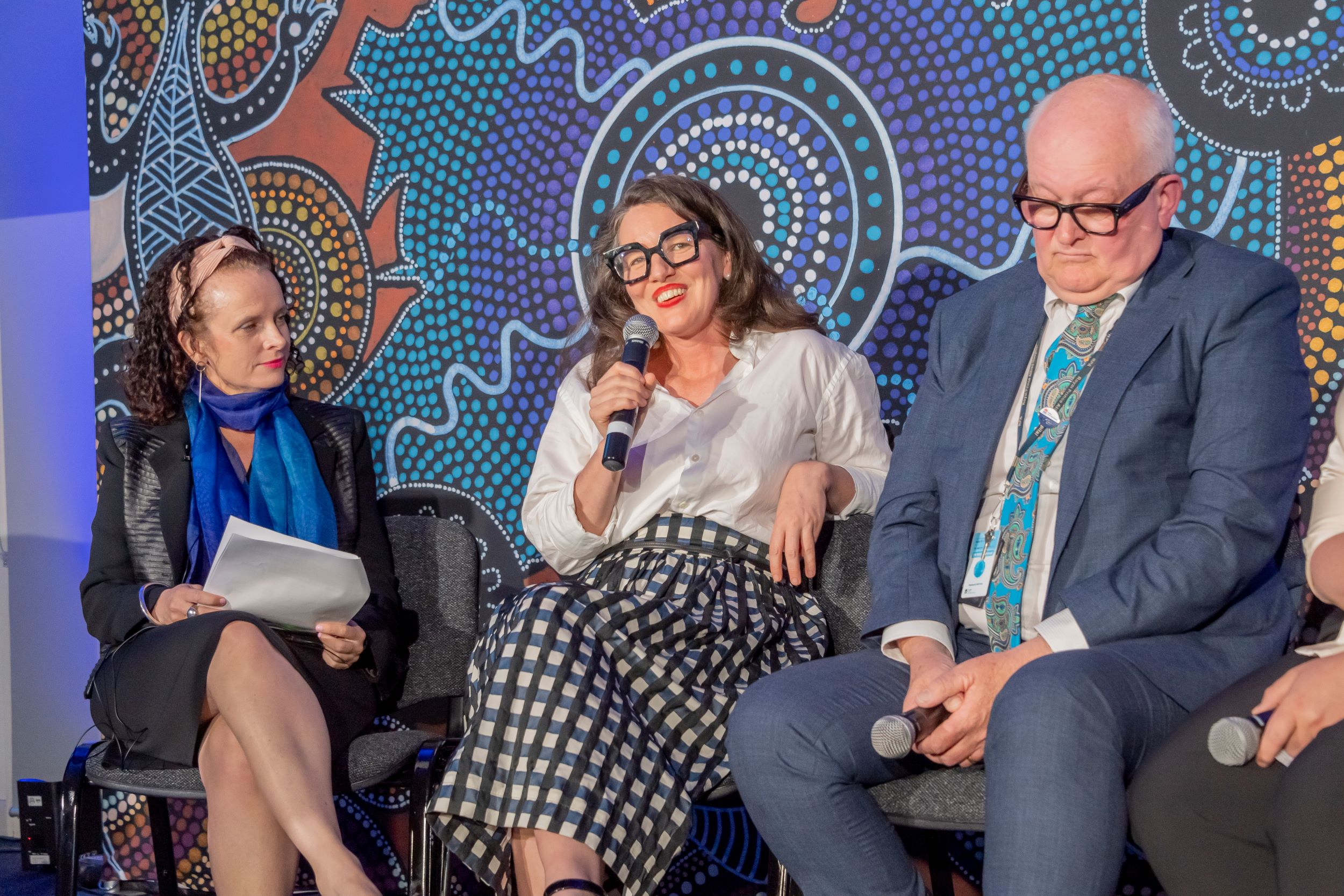 A woman sits next to another woman who is holding a microphone, and seated on the other side of her is a man.