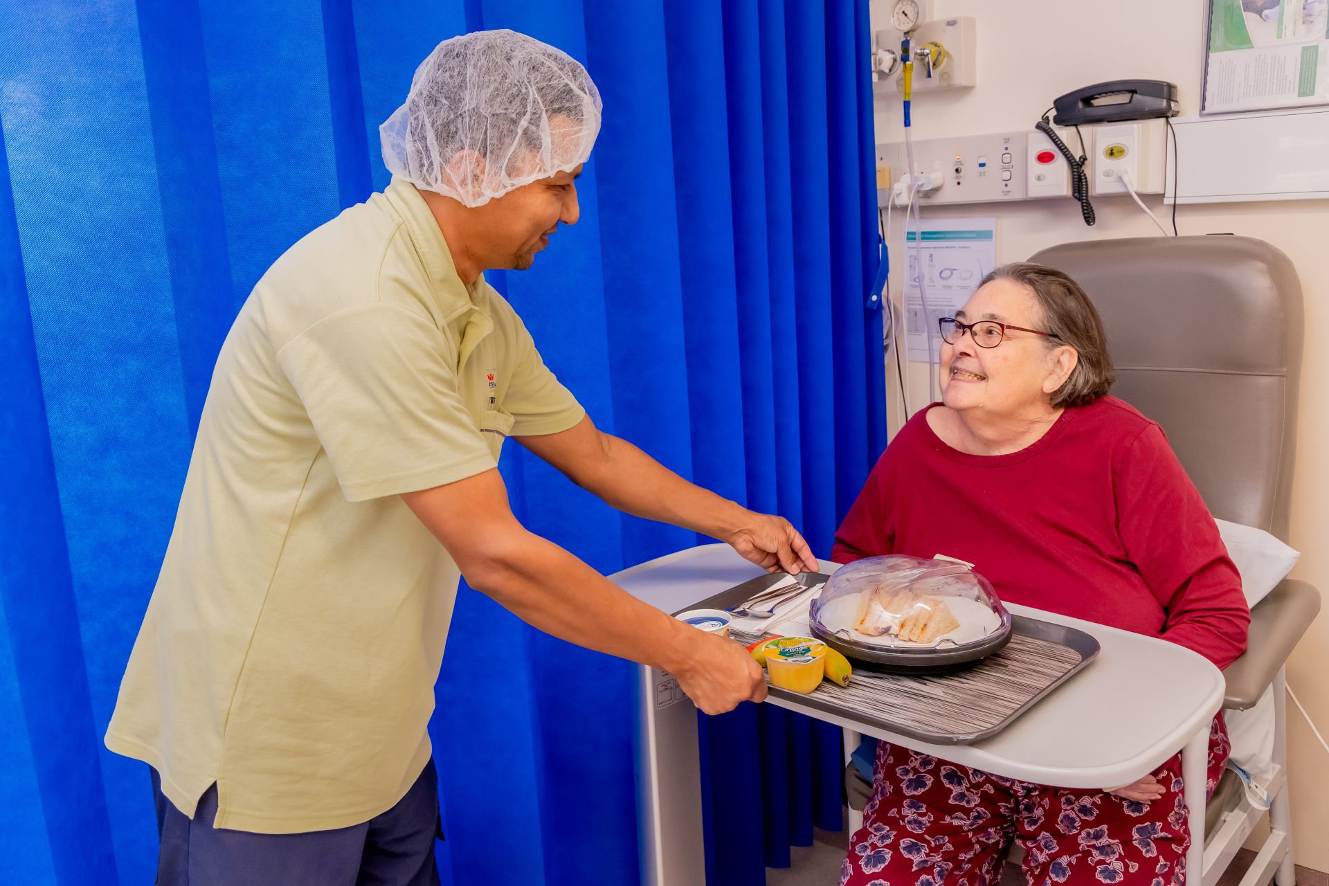 A hospital staff member delivers a food tray to a patient sitting in a chair