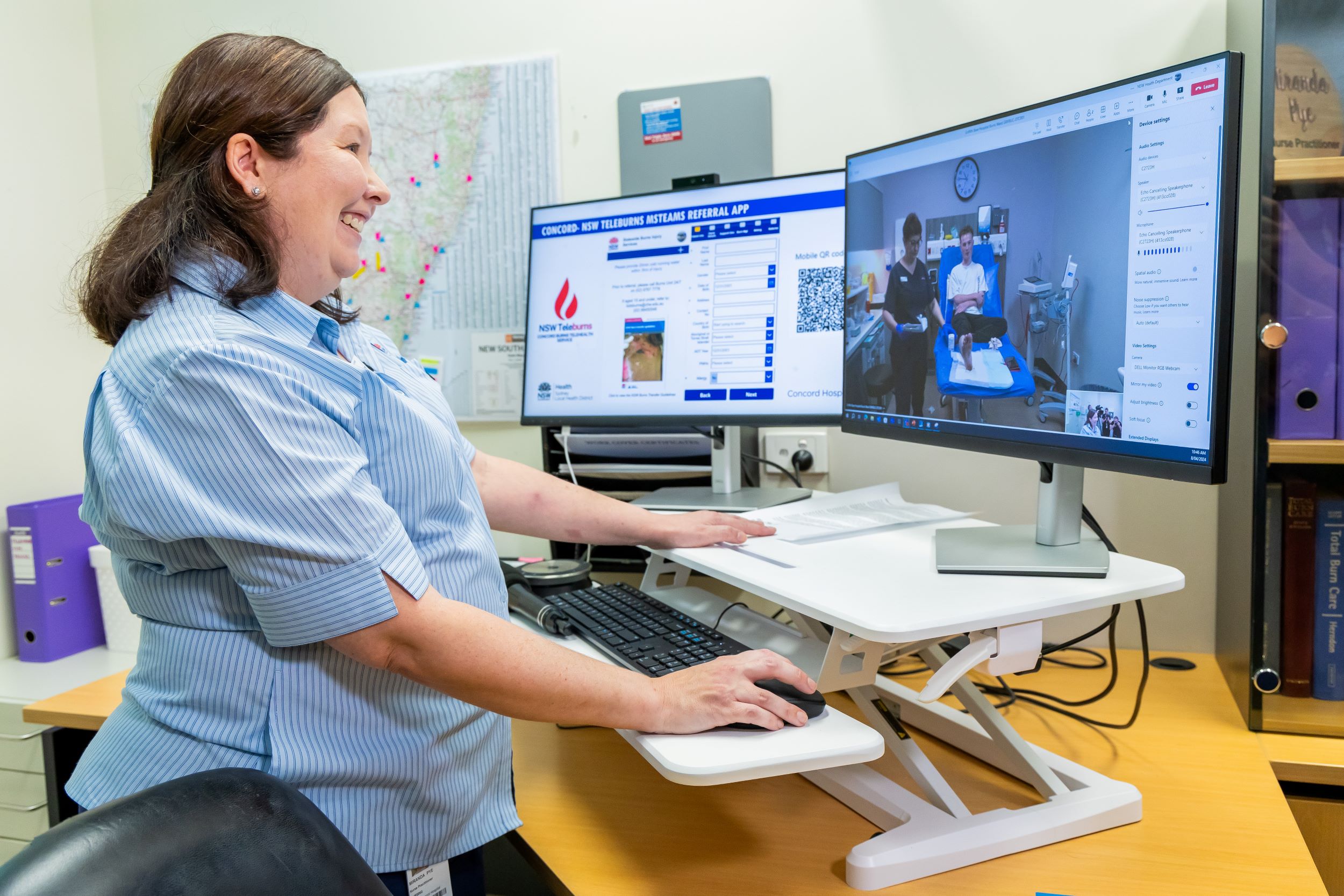 A nurse stands at a desk looking at a computer screen treating a patient in hospital in a remote location. 