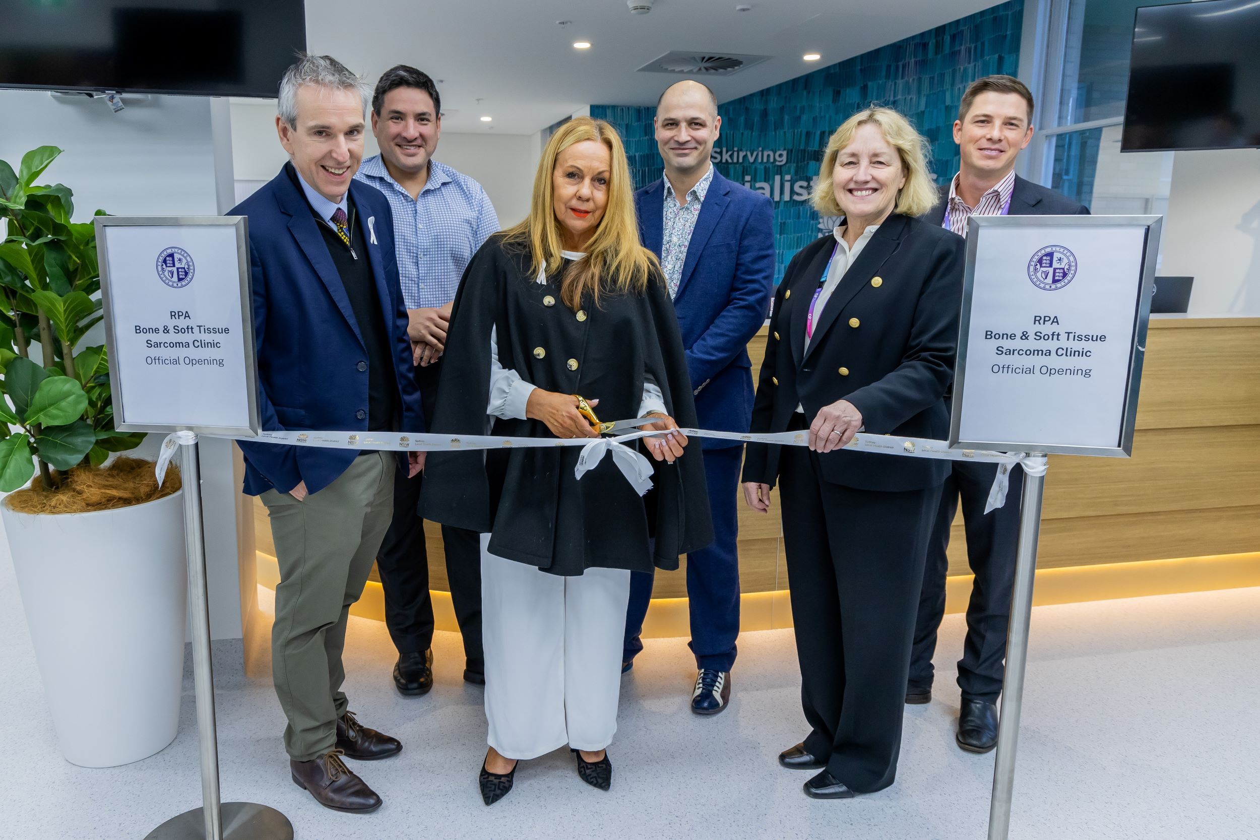 Six people stand behind a ribbon ready to cut it for the opening of a clinic.