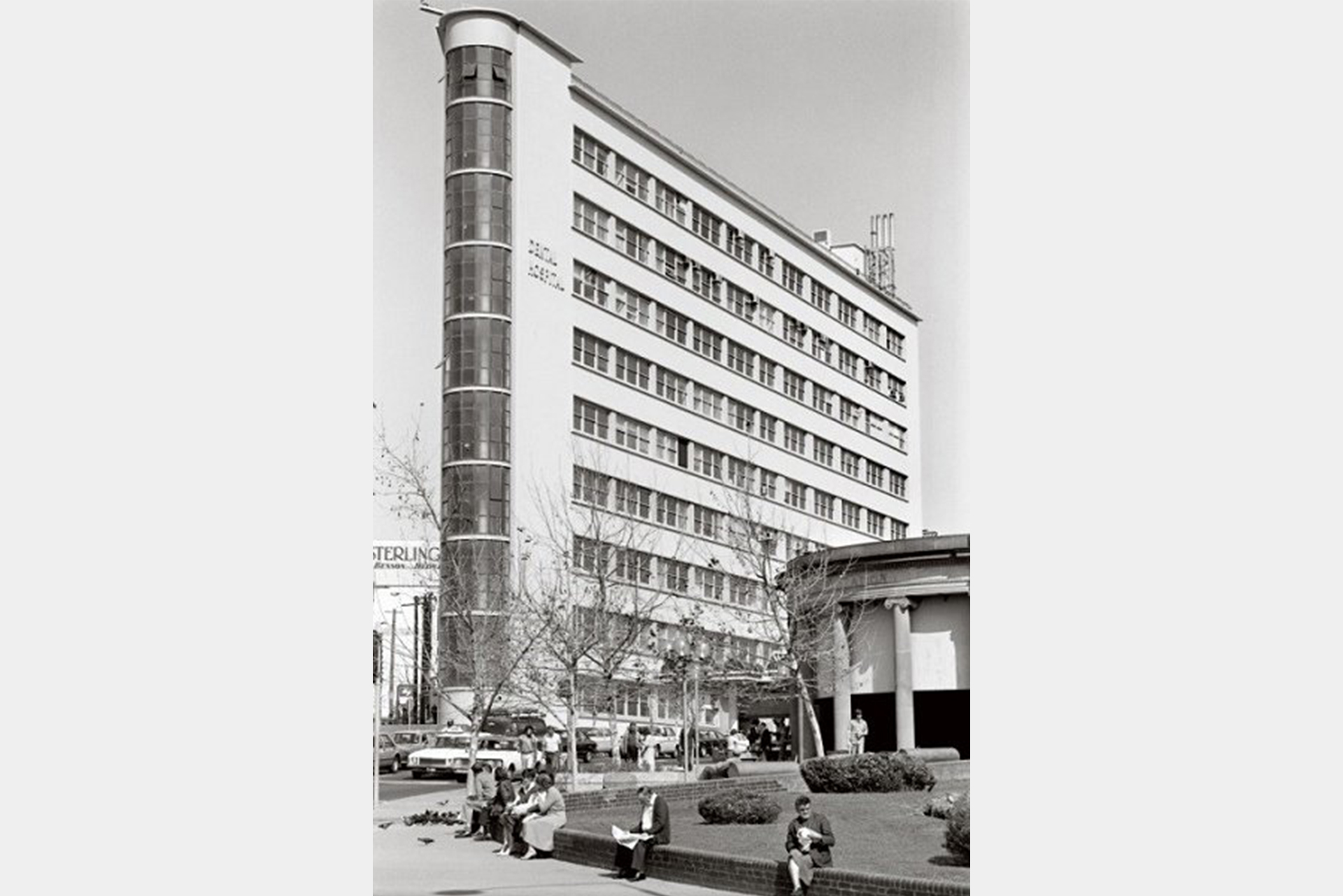 Black and white image of Sydney Dental Hospital with the entrance to Central Station's Northern Concourse and people eating lunch around the grass in the foreground.