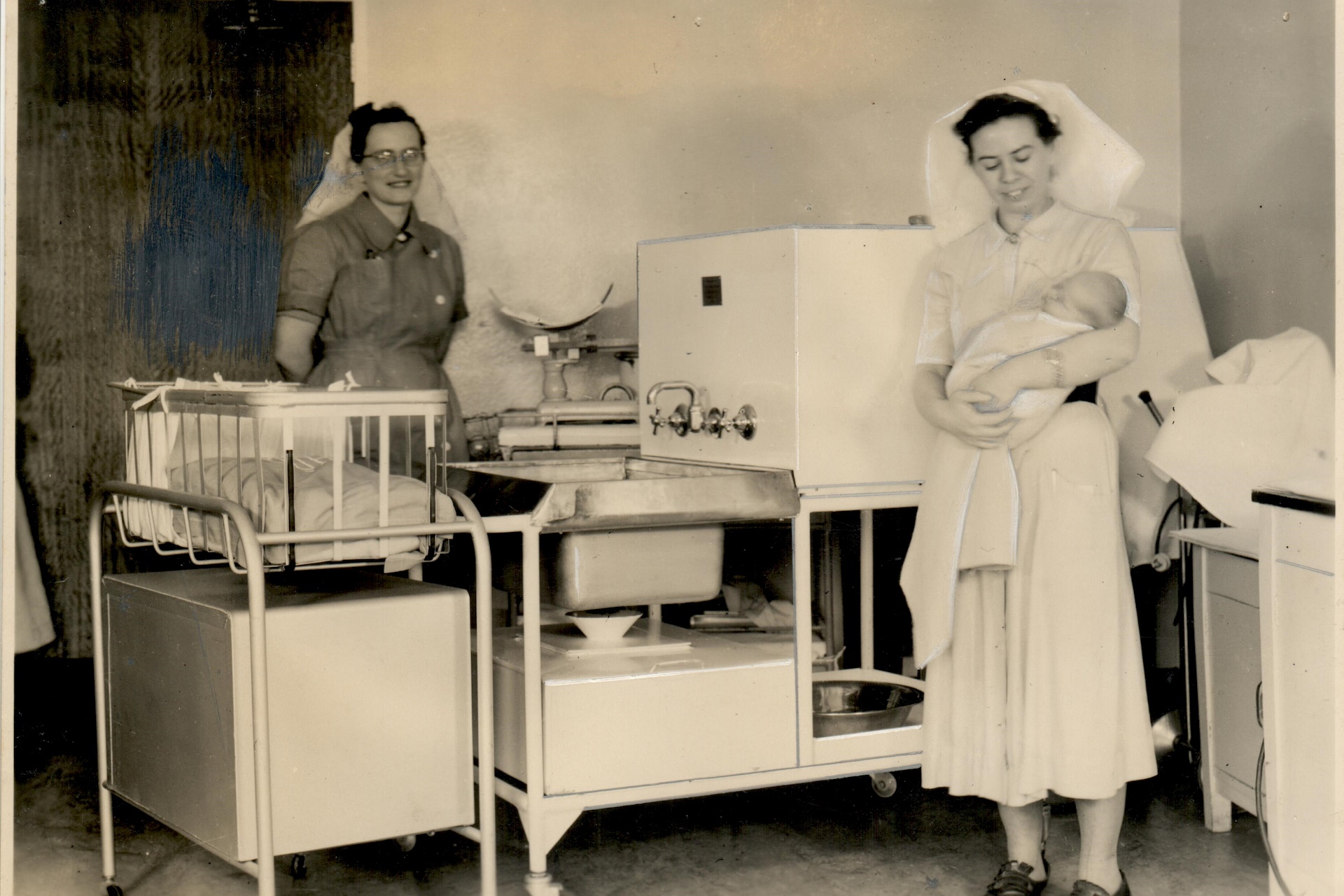 Two nurses, one holding a baby, inside a nursery in 1940