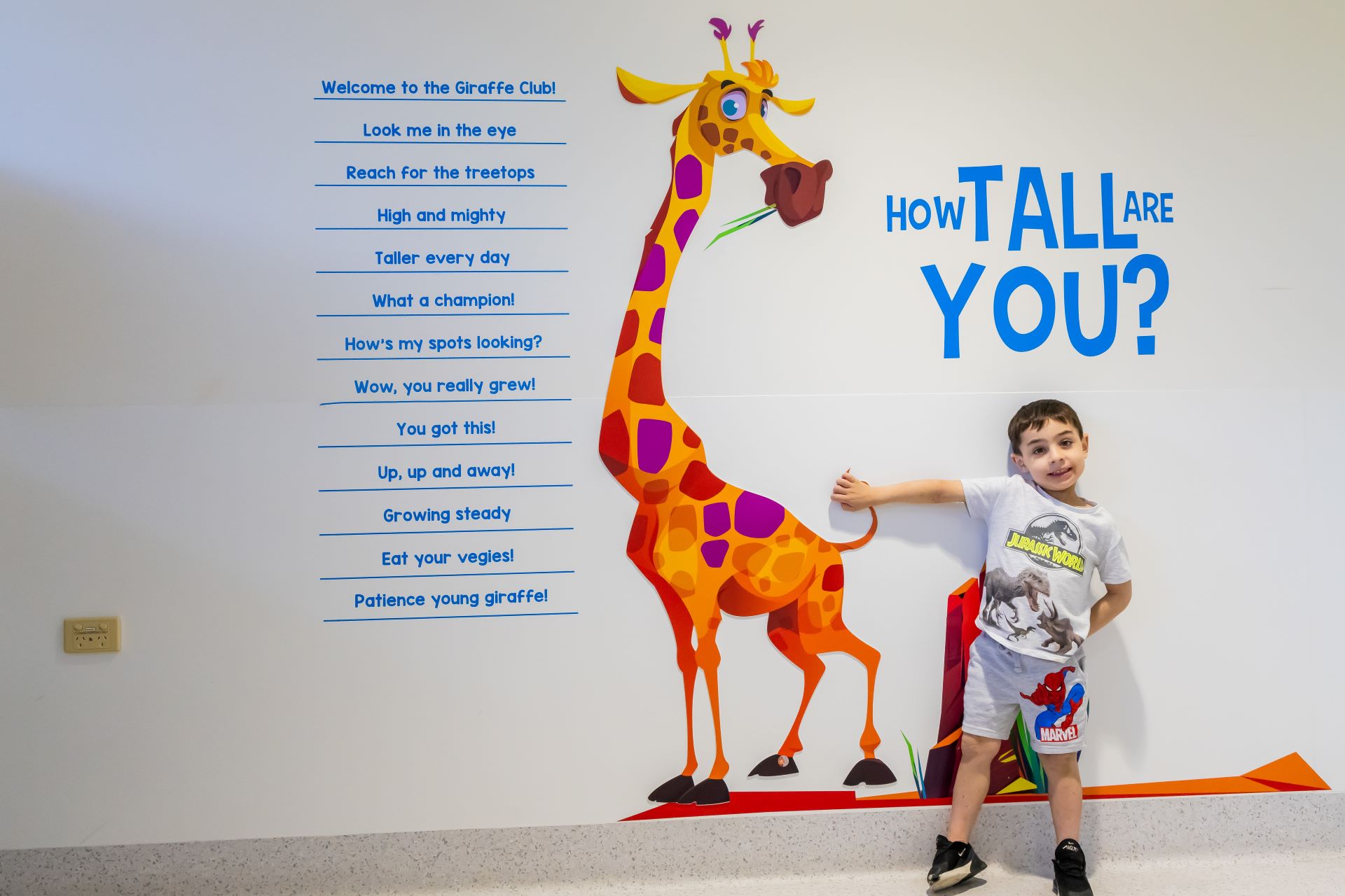 A yong boy stands next to a picture of a giraffe on a wall