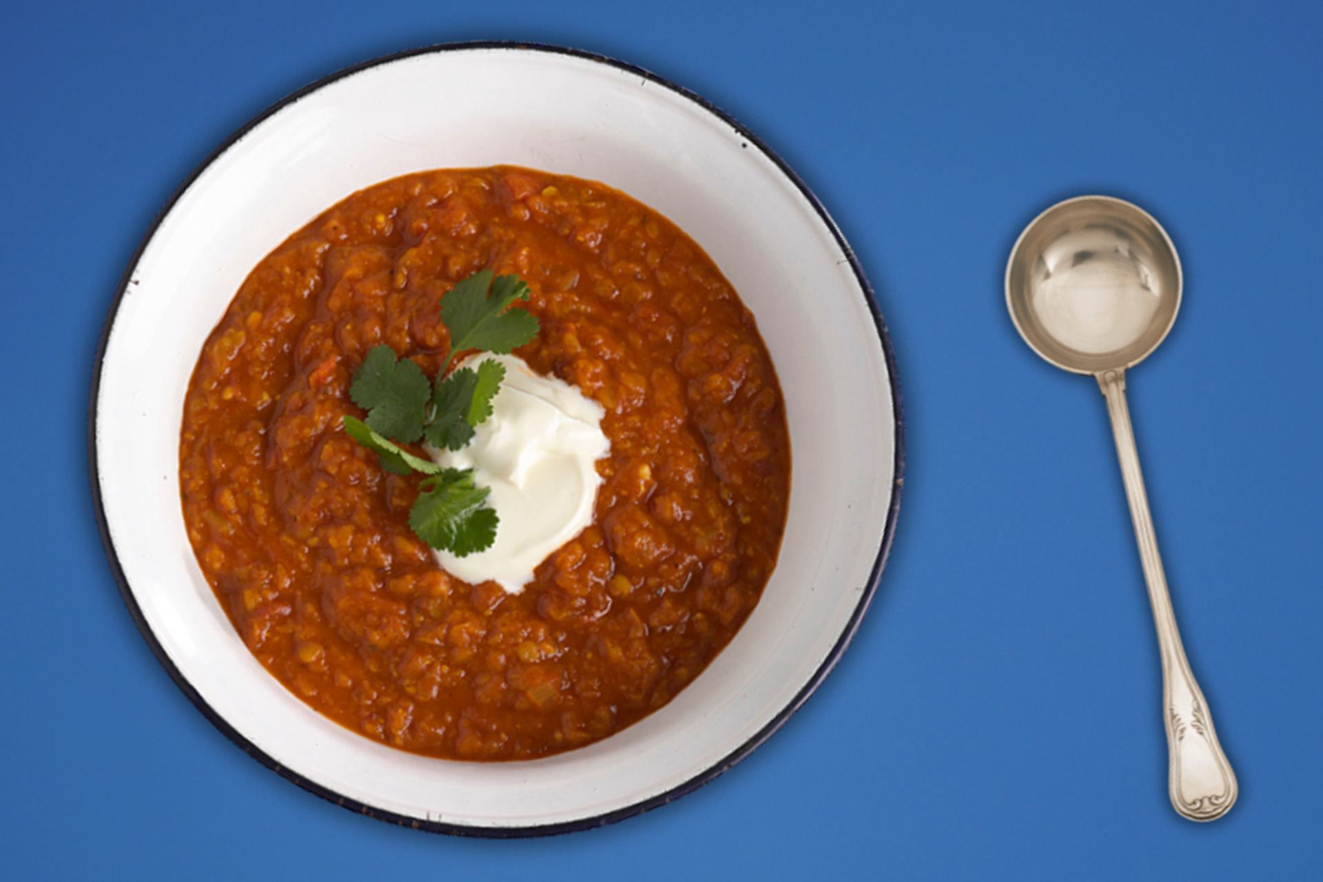 Tomato, chilli and lentil soup served in white bowl with spoon on blue table