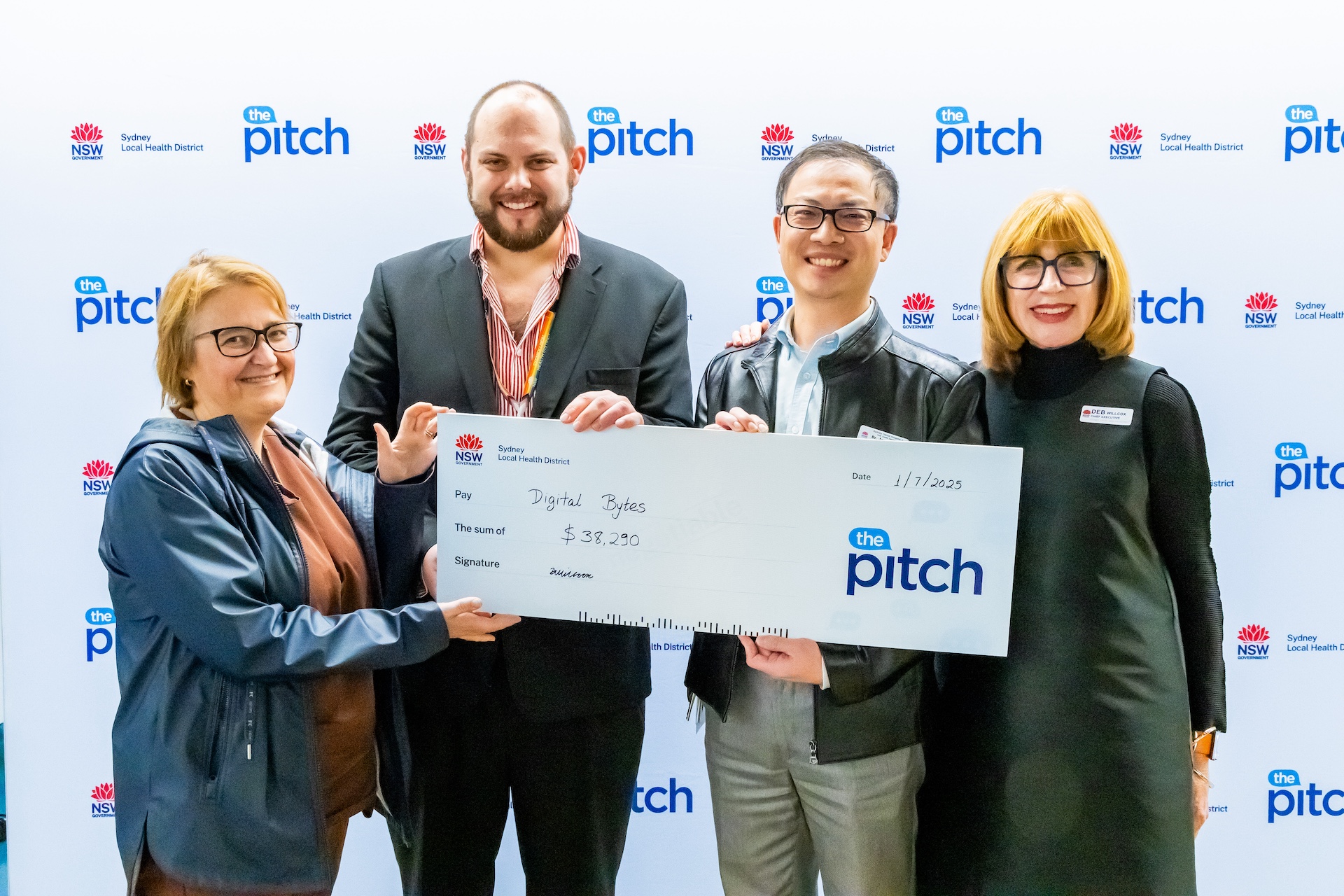 Photo of the team from Sydney Dental Hospital with Chief Executive, posing in front of the media wall with The Pitch and Sydney Local Health District logos. The staff from Sydney Dental Hospital holds a novelty check. 