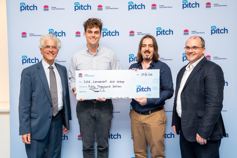 Four men standing together in front of a media wall; two in the middle holding a giant cheque