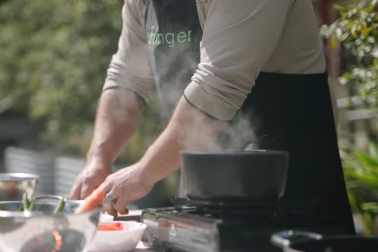 A man wearing a black apron stands next to an outdoor cooktop, preparing food