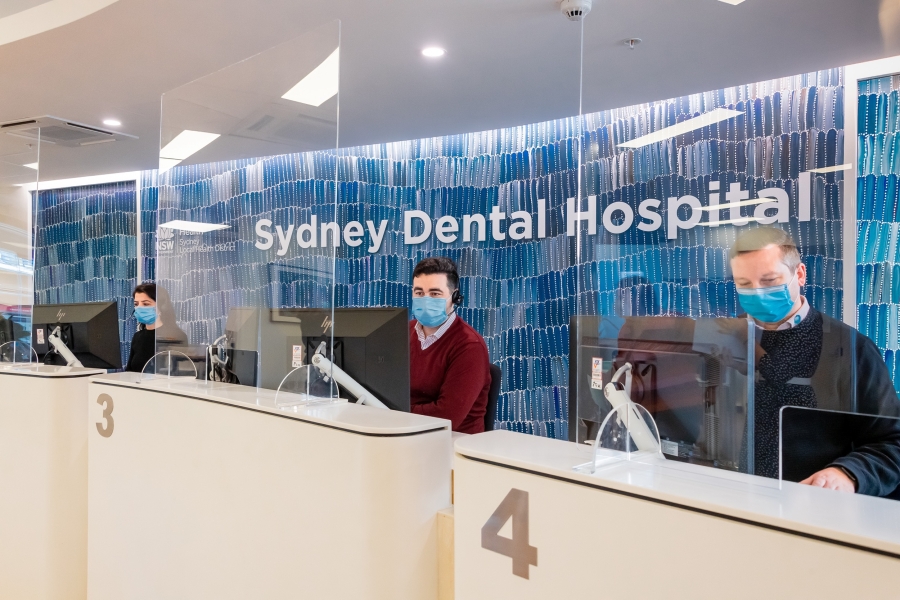 Three receptionists in front of a sign reading: ‘Sydney Dental Hospital’, which is laid on a wall of blue and white dot paintings.