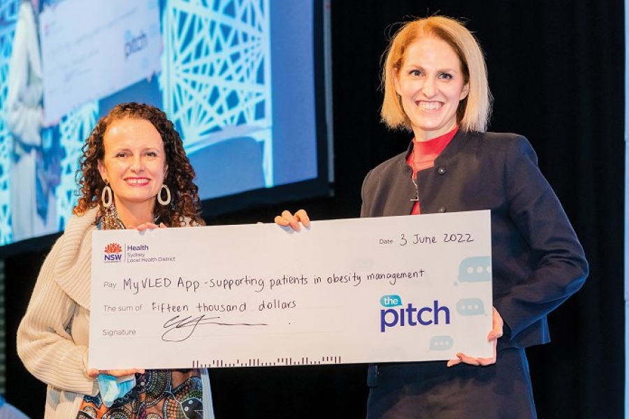 Two people hold a giant cheque reading: ‘The Pitch. Pay MyVLED App (supporting patients in obesity management) the sum of fifteen thousand dollars.