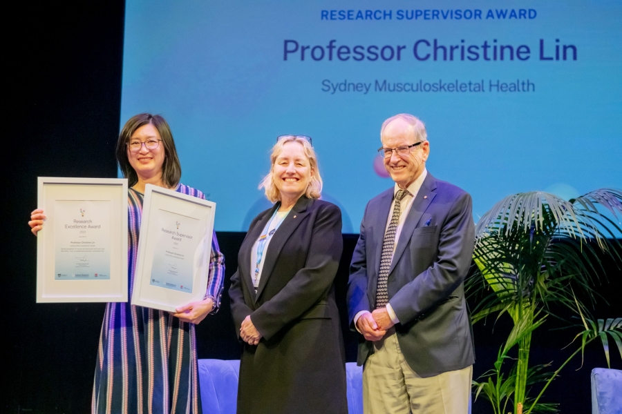 Three people standing on a stage together; first woman holding certificates