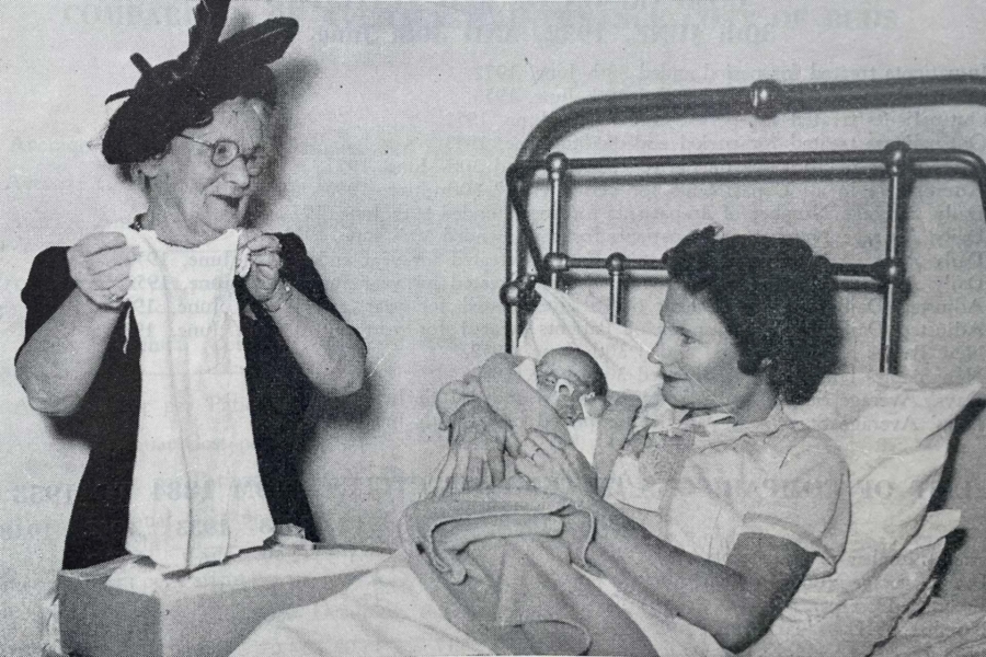 Black and white photo of a woman in bed holding her new baby with her mum on the side holding baby clothes 1953