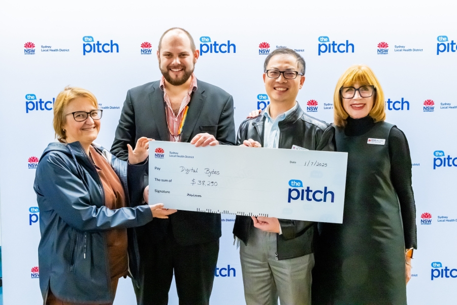 Photo of the team from Sydney Dental Hospital with Chief Executive, posing in front of the media wall with The Pitch and Sydney Local Health District logos. The staff from Sydney Dental Hospital holds a novelty check. 